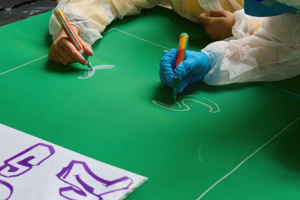 Zwei Kinderhände malen mit Markern den Letter S auf ein grün bemaltes Holzbrett.