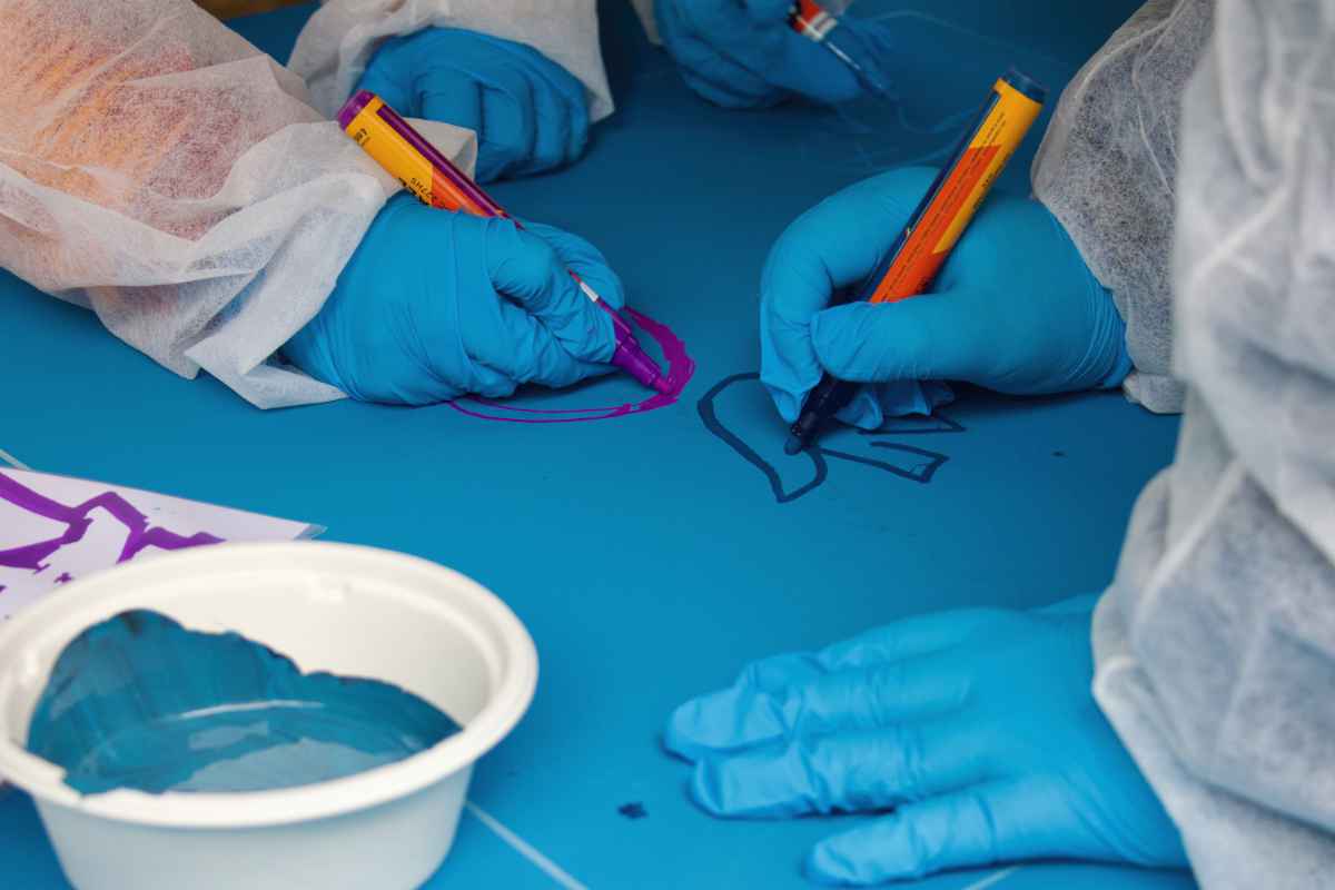 Mehrere Kinderhände in Handschuhen malen mit Markern auf ein blau bemaltes Holzbrett.