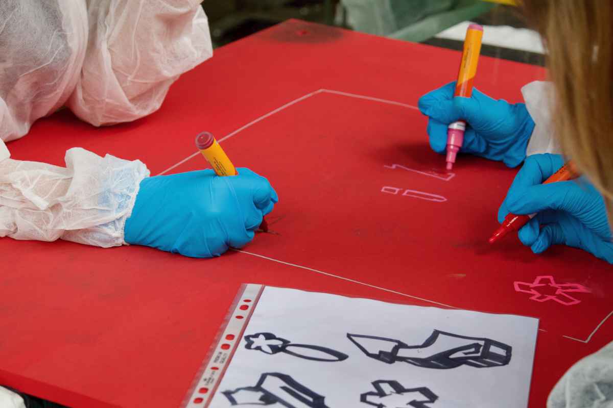 Drei Kinderhände in Handschuhen malen mit Markern auf ein rot bemaltes Holzbrett den Letter i und Sterne.