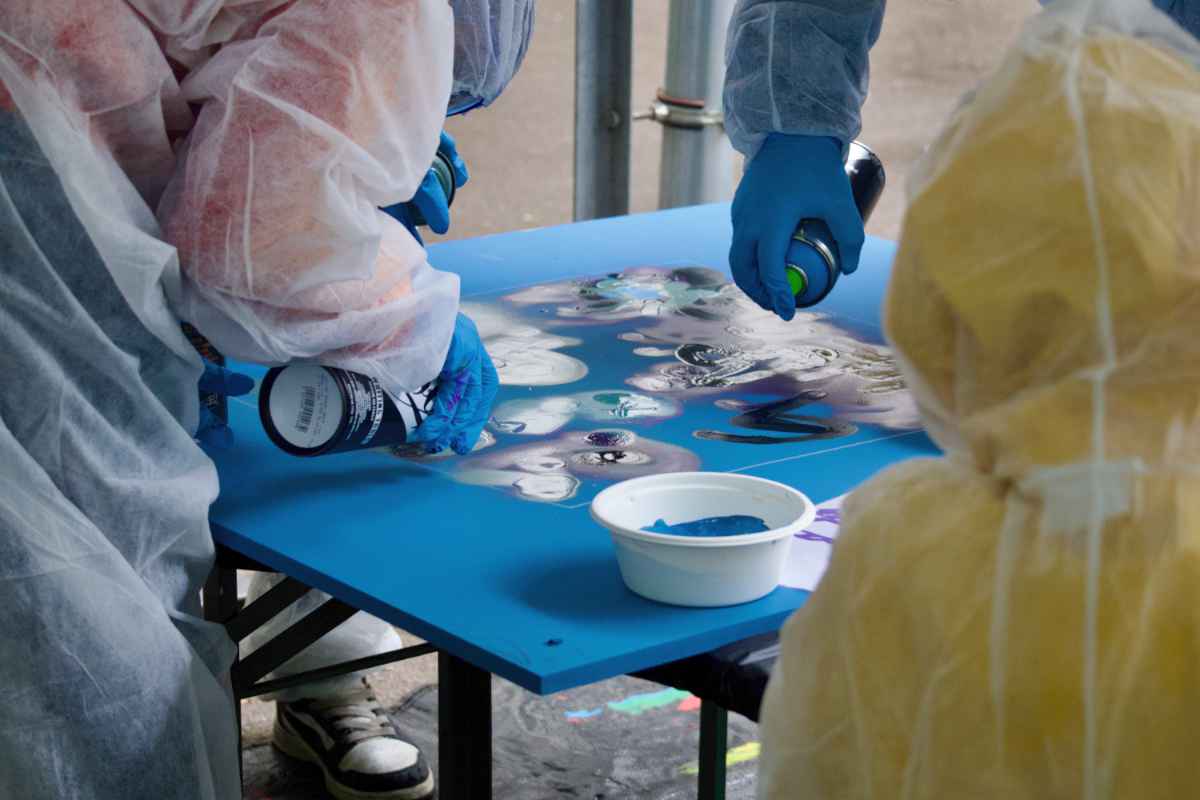 Mehrere Kinder in Schutzausrüstung sprayen Graffiti auf eine liegende blaue Holzplatte. Es ist nicht zu erkennen was gesprüht wird.
