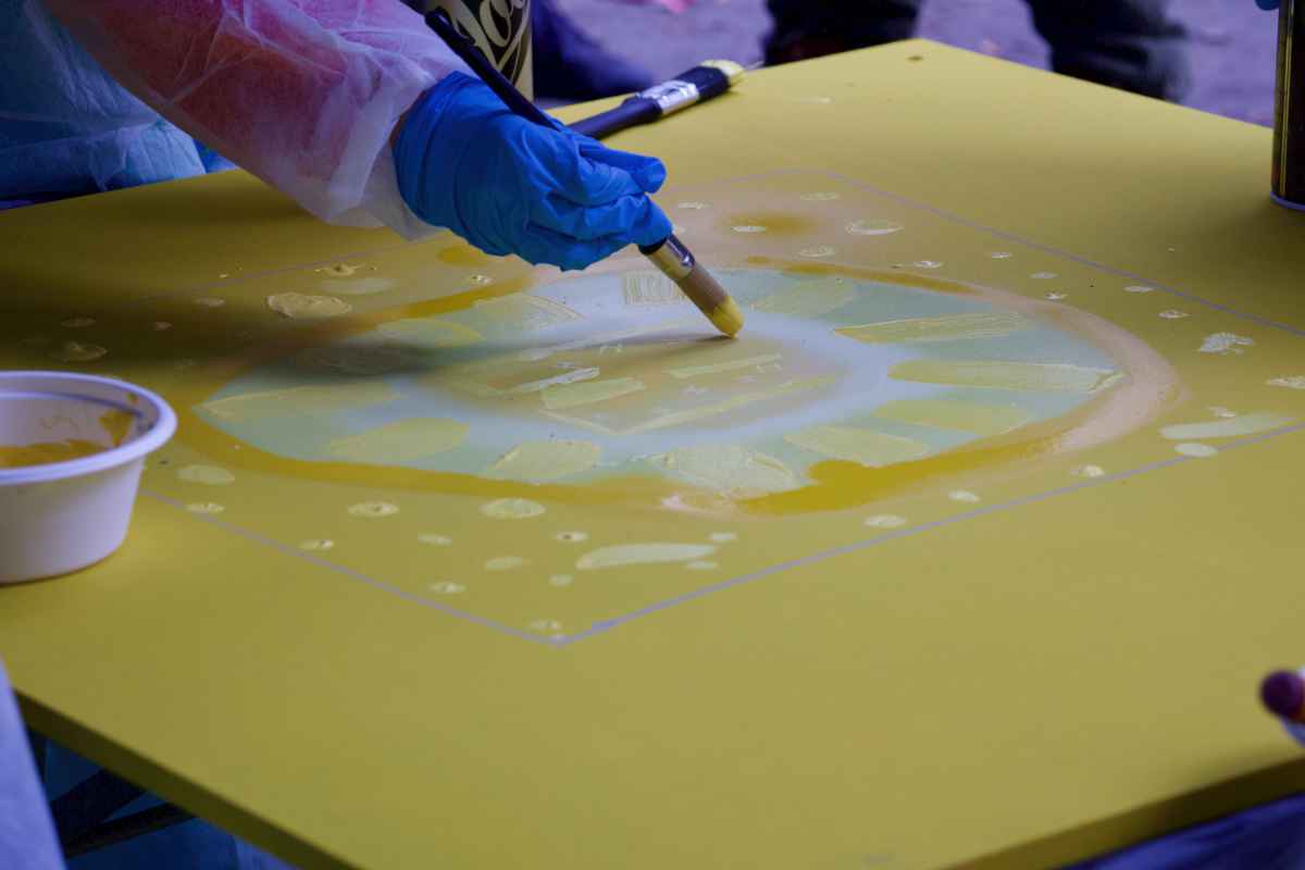 Ein Kind mit Handschuhen malt mit einem dicken Pinsel ein gelbes H auf eine liegende gelbe Holzplatte.