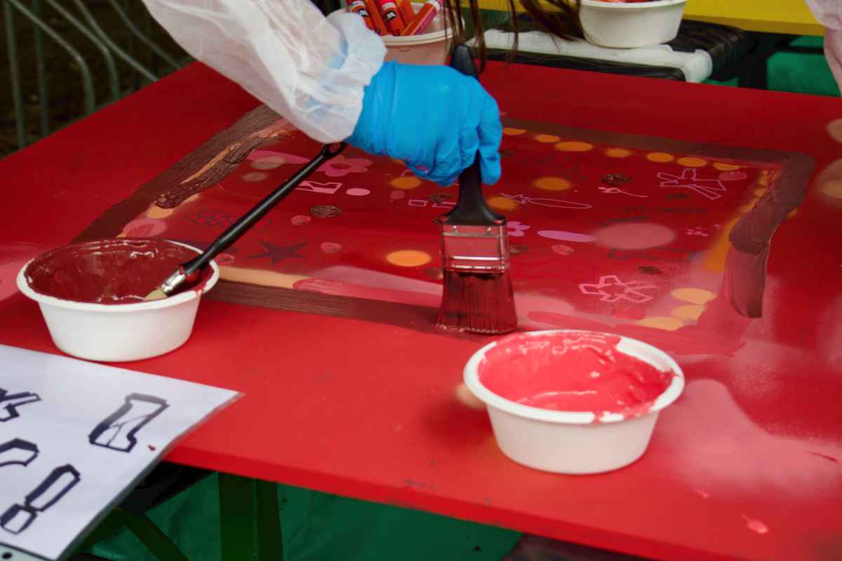 Ein Kind in Schutzmontur streicht mit einem breiten Pinsel dunkelrote Farbe auf eine rote Holzplatte.