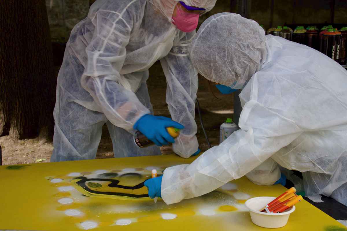 Ein Kind sprüht gelbe Farbe auf eine gelbe Holzplatte, während ein zweites Kind mit einem gelben Marker auf dieselbe Platte malt. Beide tragen Schutzausrüstung.