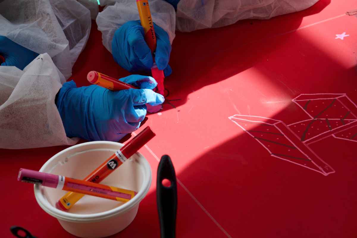 Zwei Kinder malen gemeinsam an einer detaillierten Grafik mit roten Markern auf eine rote Holzplatte. Auf der Platte ist bereits eine 3D-Kontur des Buchstaben H gemalt.