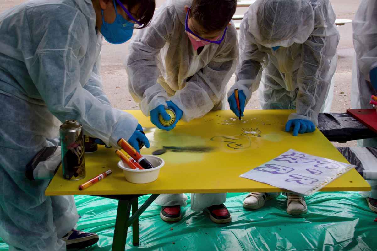 Drei Kinder bemalen in Zusammenarbeit eine gelbe Holzplatte. Zwei mit Sprühfarbe und eins mit einem Marker.