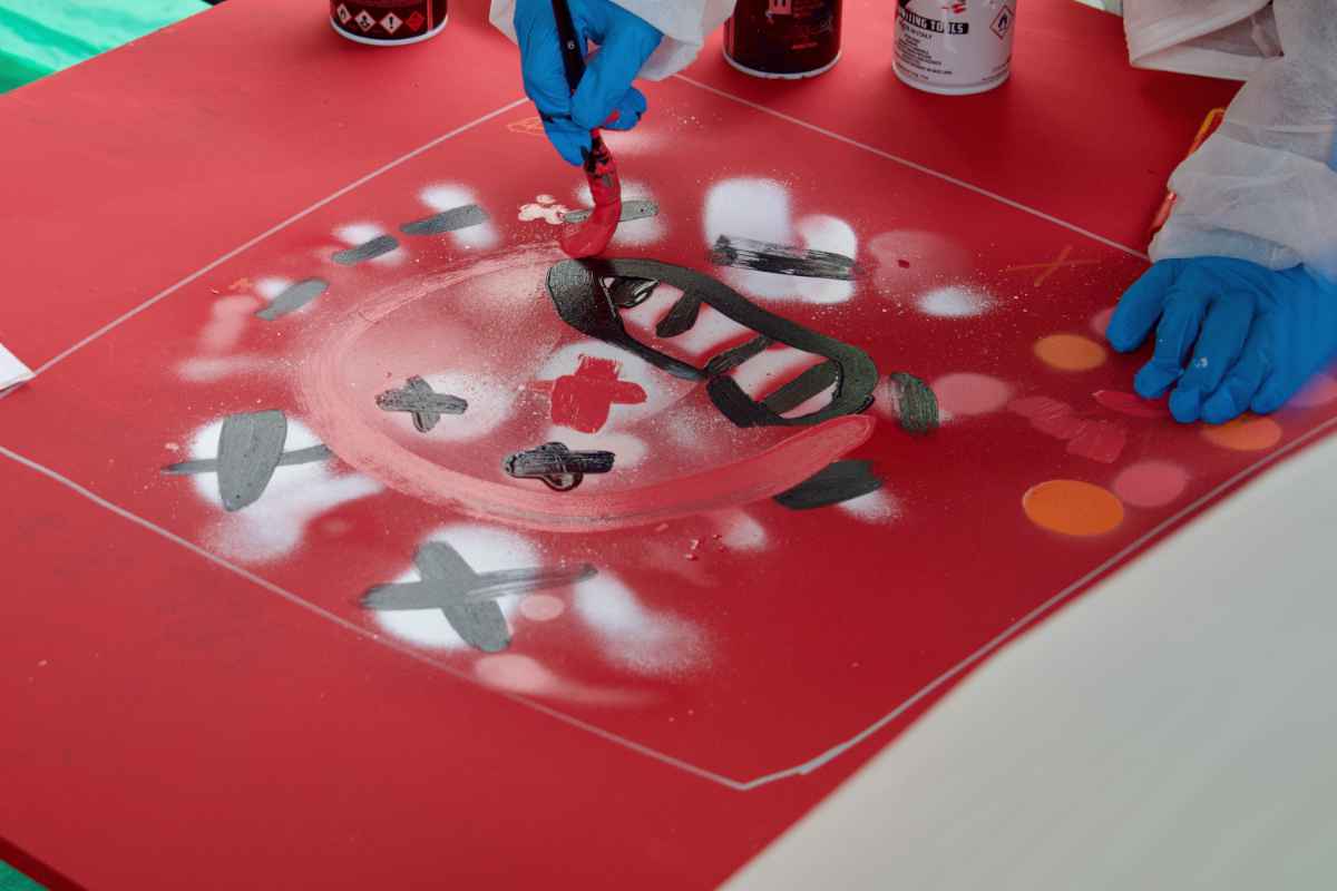 Die Kontur eines Charakters wird mittels Pinsel und roter Farbe von einem Kind auf eine rote Holzplatte gemalt. Man erkennt bereits ein gemaltes Gesicht.