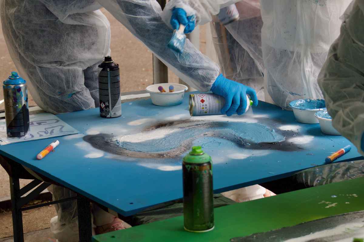 Ein Kind srpüht weiße Farbe auf eine blaue Holzplatte. Die Kinder tragen wie immer Schutzausrüstung. Weitere Marker und Sprühdosen stehen auf der Platte.
