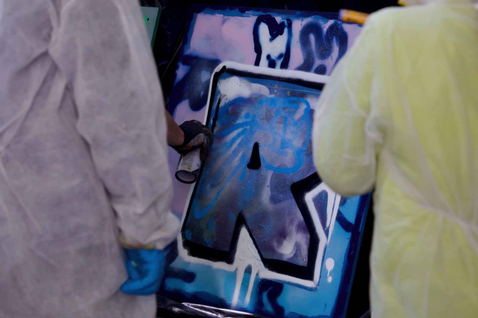 Die Hand eines Graffiti-Sprayers zeichnet die Kontur des Letters R auf einem Holzbrett nach. Im Vordergrund die Rückseite zweier Kinder in kompletter Schutzausrüstung.