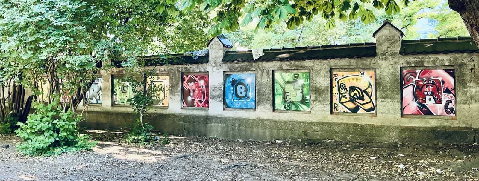 Steinmauer auf dem Hof der Hirschbergschule. An der Mauer sind 10 große mit Graffiti bemalte Holzplatten montiert. Jede Platte zeigt einen Buchstaben. Die Buchstaben sind H, I, R, S, C, H, B, E, R und G. Zusammengelesen erbigt das Hirschberg
