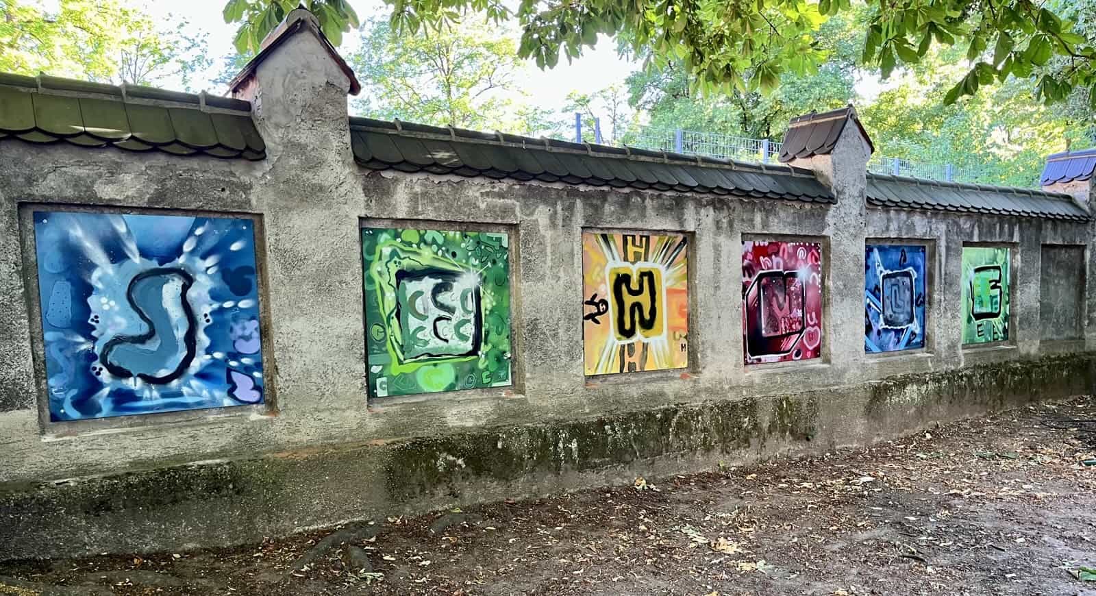Das Endergebnis der Kinder ist auf der Steinmauer auf dem Hof der Hirschbergschule zu sehen. Das Bild zeigt auf die Mauer montierte Holzplatten, auf die jeweils mit Graffiti gemalt wurde. Auf jeder Platte ist ein Buchstabe zu sehen. S, C, H, U, L und E. Zusammengelesen erbigt das Schule. Alle Platten verfolgen ein einheitliches Farbschema. Die Platten sind in rot, gelb, grün und Blautönen gehalten.